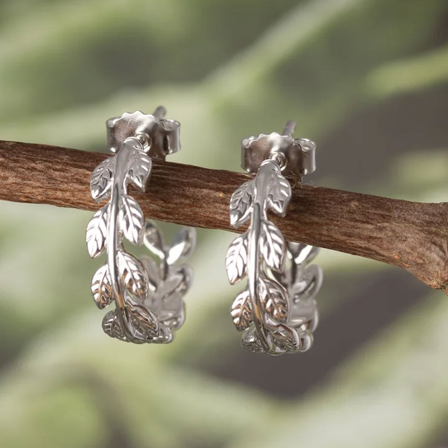 A pair of silver huggie hoop earrings with detailed leaf and vine motifs, displayed on a small brown twig against a soft, blurred green background.