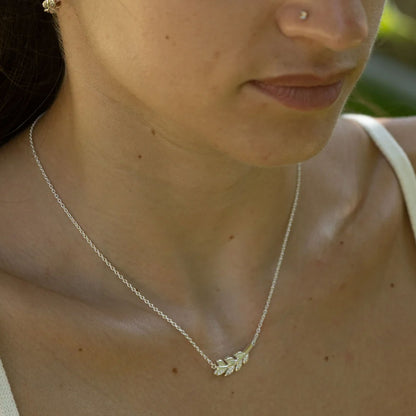 A close-up lifestyle shot of a woman wearing a delicate silver chain necklace with a small leaf-shaped pendant, paired with a white tank top.