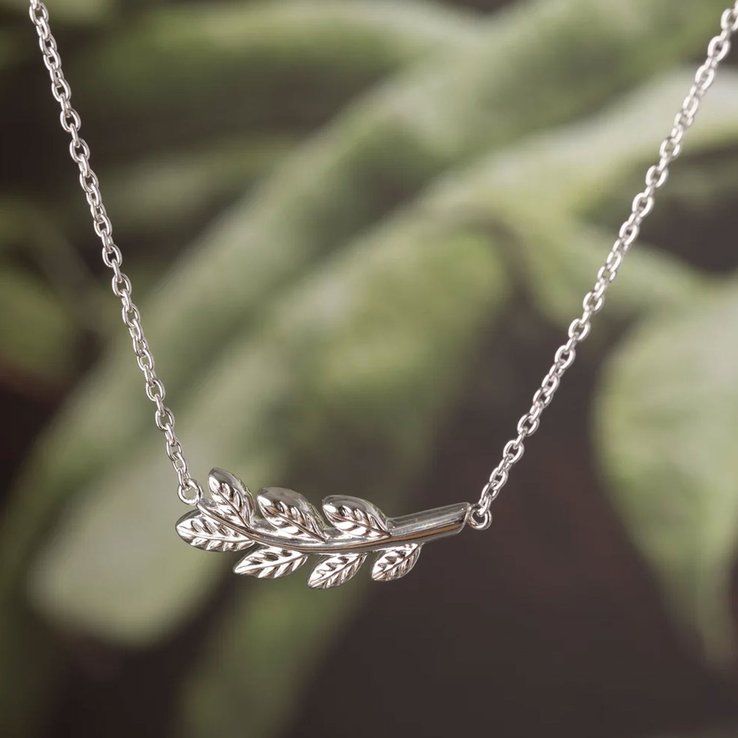 A silver laurel leaf branch pendant necklace on a delicate silver chain, shown against a soft, blurred green background.