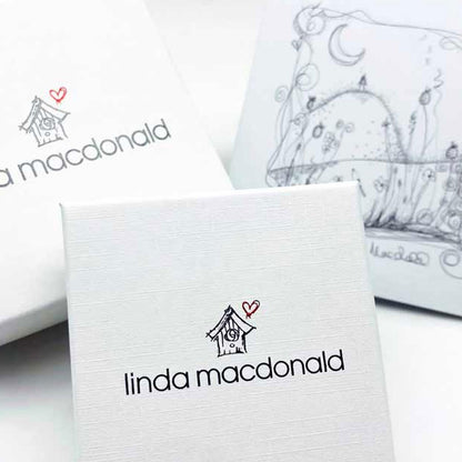 A close-up of a white textured gift box printed with the name 'linda macdonald' and a small house-with-heart logo, with additional branded packaging and a sketchy illustration visible in the background.