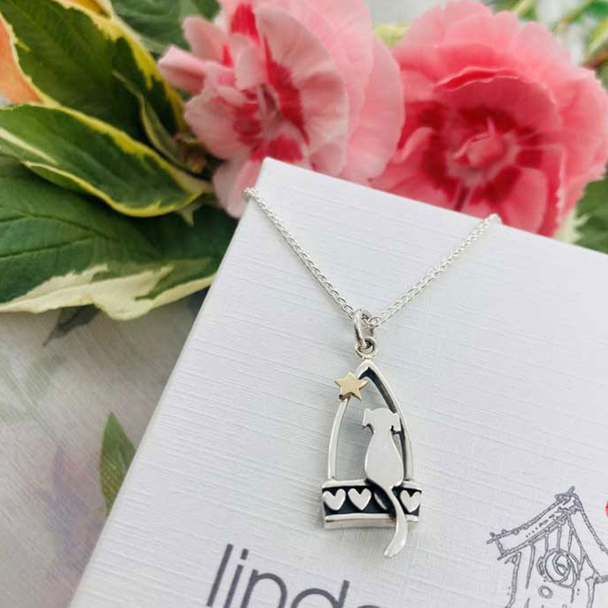 A silver pendant necklace featuring a dog silhouette sitting on a bench with heart details and a small gold star, displayed on a white box with pink flowers in the background.