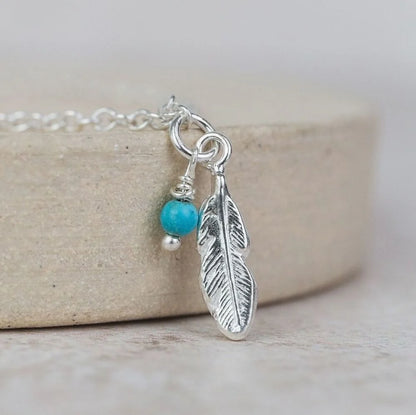 Silver feather charm with turquoise bead on a chain against a neutral background