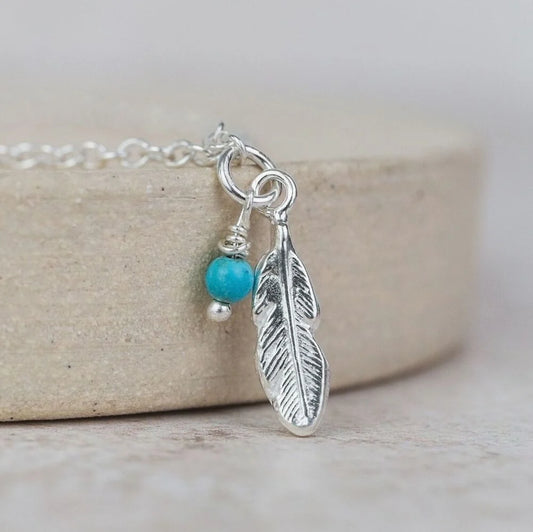 Silver feather charm with turquoise bead on a chain against a neutral background