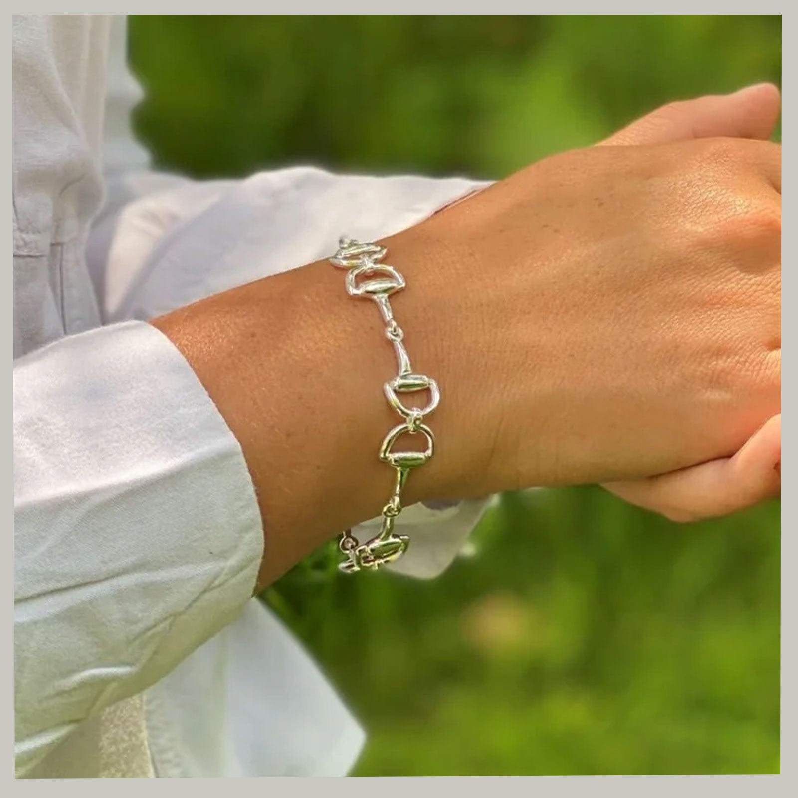 A silver horse-bit style chain bracelet worn on a person's wrist, with a white long-sleeve shirt visible and a blurred green outdoor background.