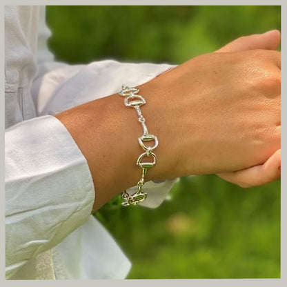 A silver horse-bit style chain bracelet worn on a person's wrist, with a white long-sleeve shirt visible and a blurred green outdoor background.