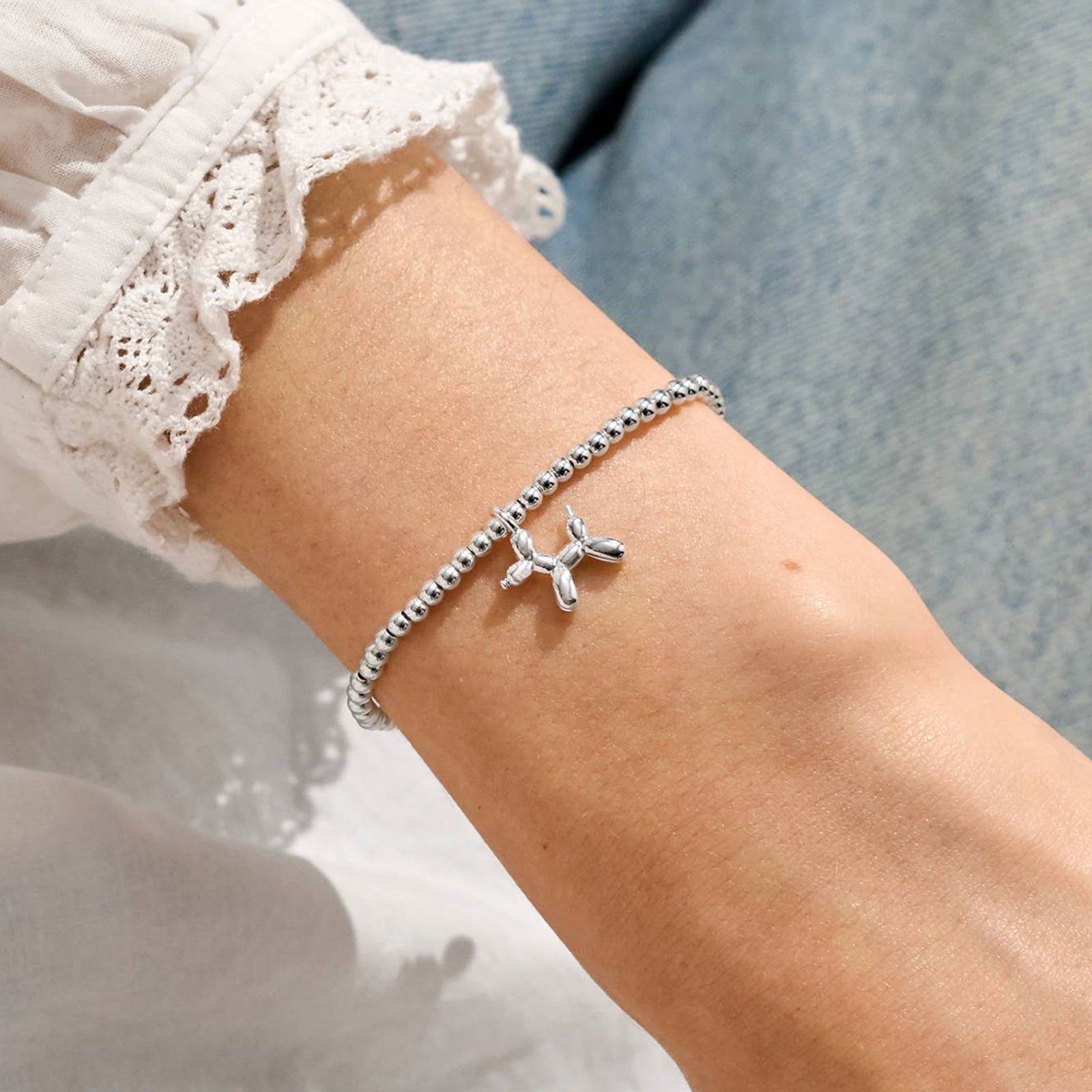 A silver beaded bracelet with a small dog-shaped charm worn on a person's wrist, paired with a white lace-sleeve blouse and blue denim in the background.