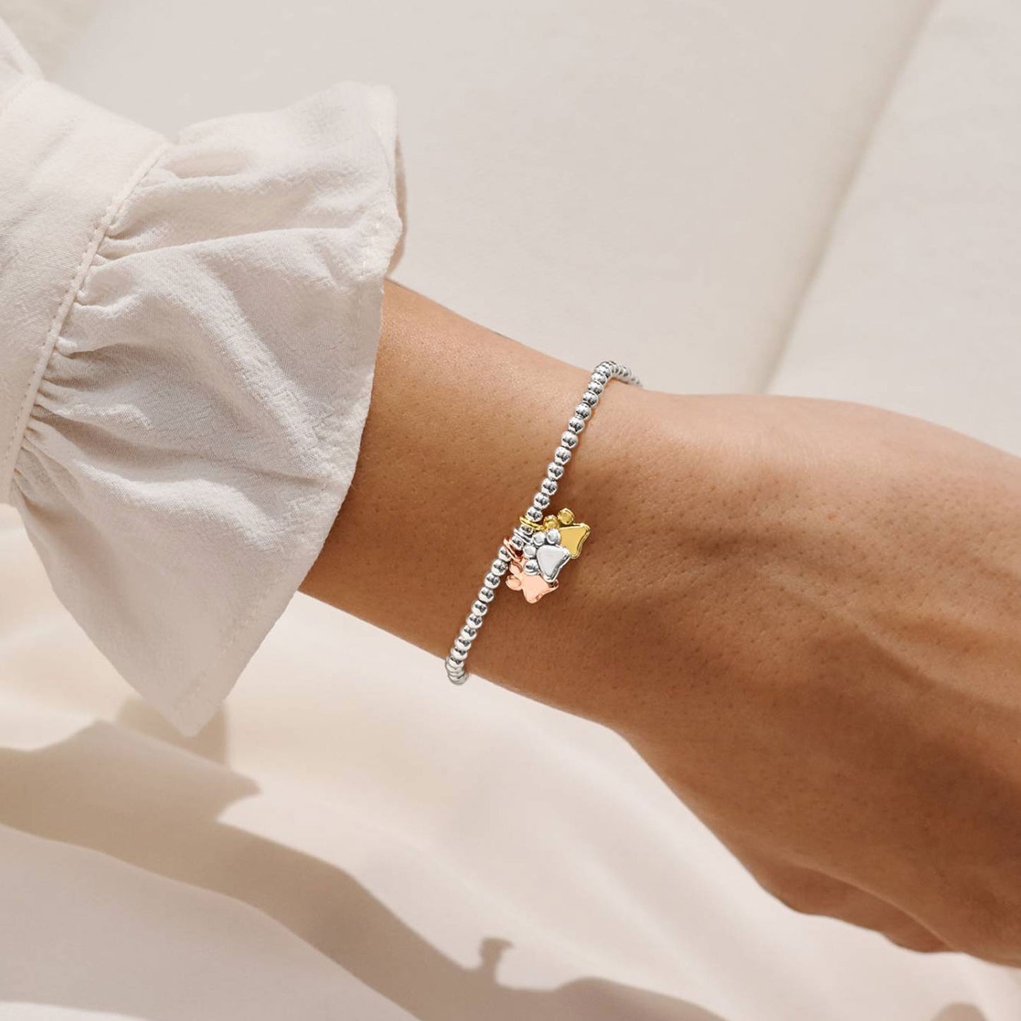 A close-up of a person's wrist wearing a delicate silver beaded bracelet with a small gold and rose gold crown charm, styled with a white ruffled sleeve.