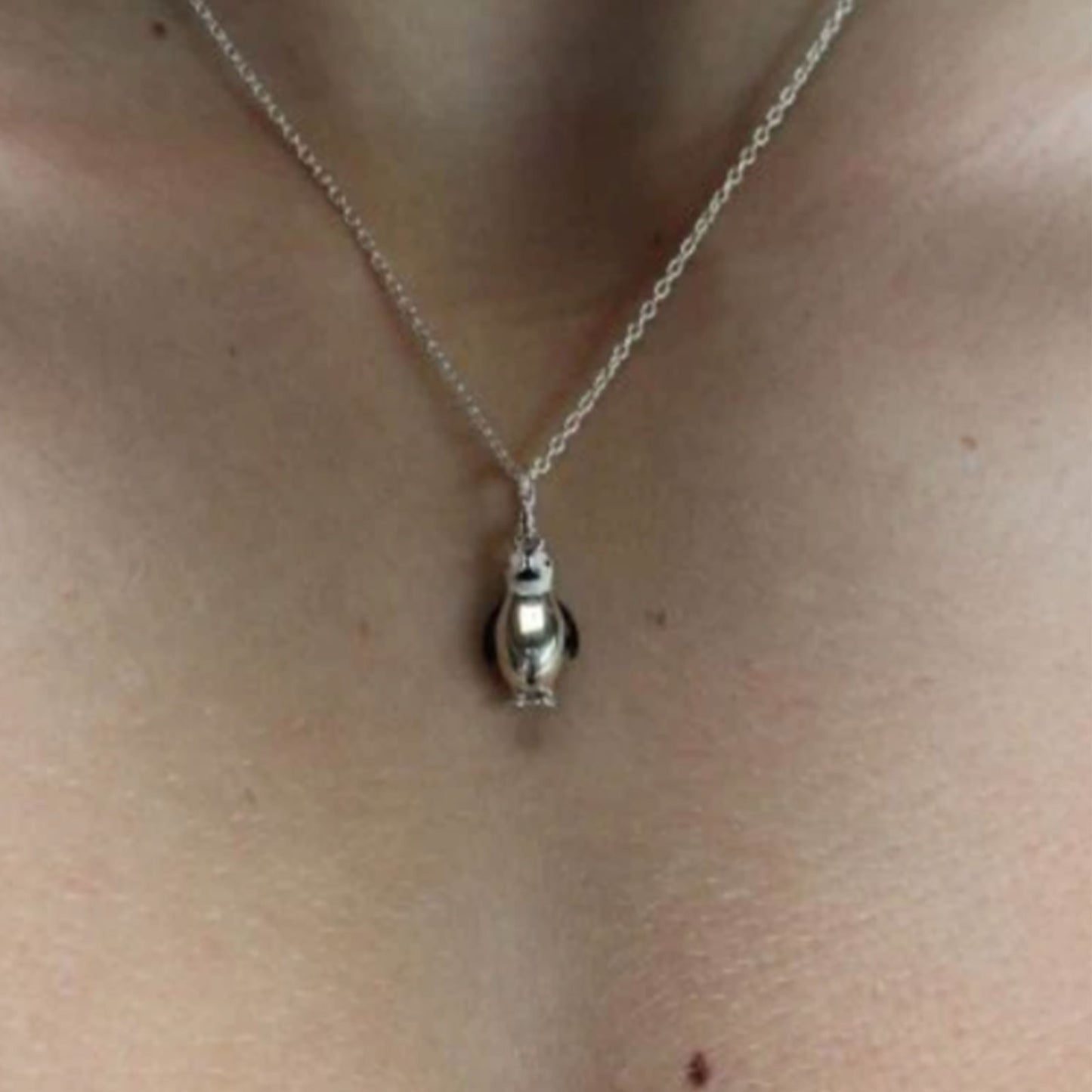 A close-up of a person's neck and chest wearing a delicate silver chain necklace with a small penguin-shaped pendant.