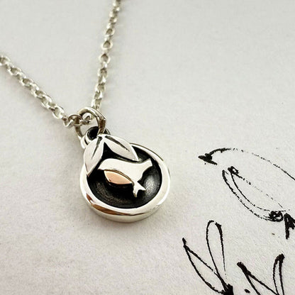 A silver pendant necklace featuring a stylized bird design over a black circular background, displayed on a textured off-white surface with a black sketch.