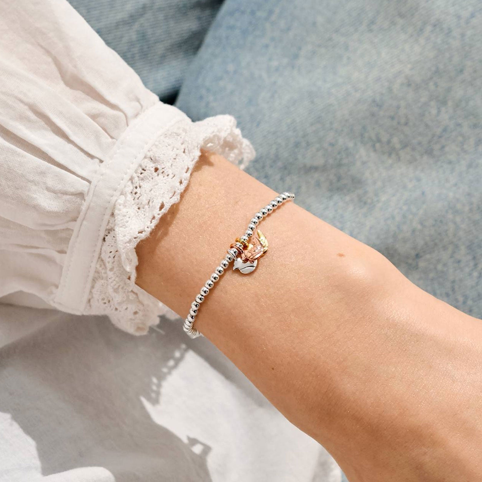 A delicate silver beaded bracelet with a small charm featuring a pink and yellow butterfly, worn on a person's wrist. The person is wearing a white lace-sleeve blouse and light blue denim jeans.