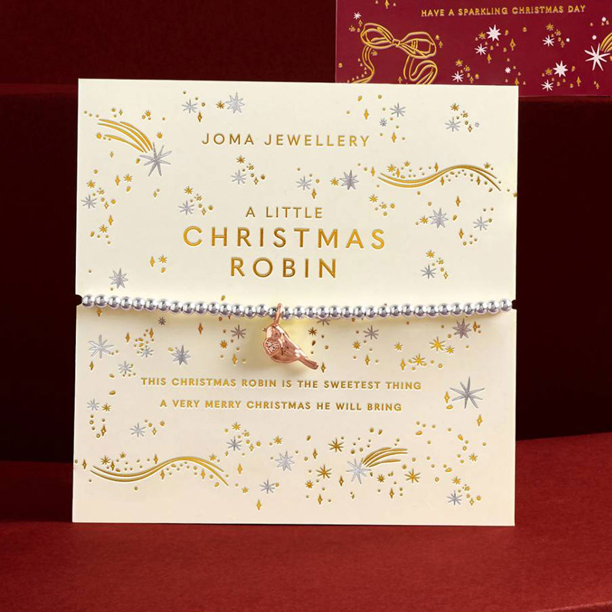 A Joma Jewellery Christmas-themed beaded bracelet with a small robin bird charm, displayed on its cream-colored branded card packaging against a dark red background.