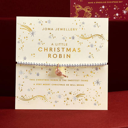 A Joma Jewellery Christmas-themed beaded bracelet with a small robin bird charm, displayed on its cream-colored branded card packaging against a dark red background.