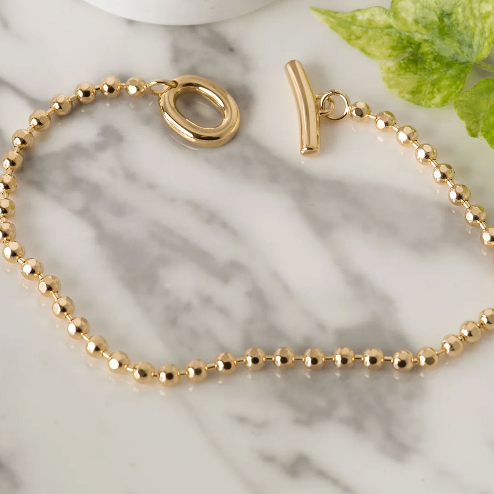 Gold bracelet with a clasp on a marble surface with a green leaf.