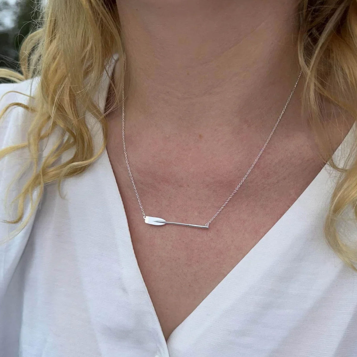 A close-up of a woman wearing a delicate silver necklace with a rowing blade / oar, styled with a white blouse.