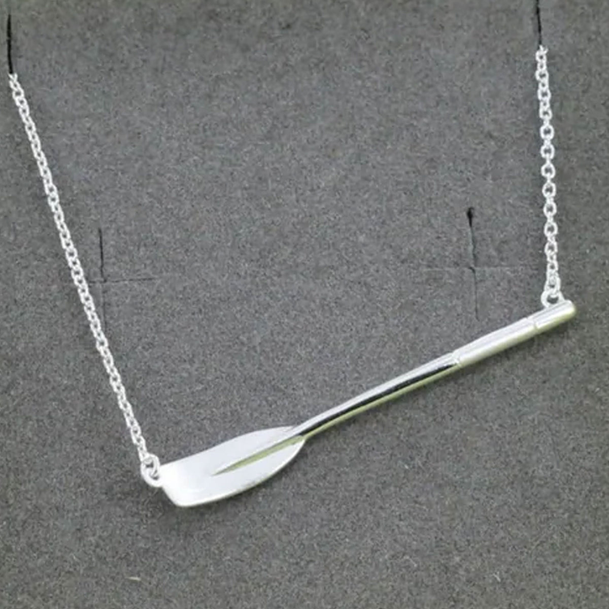 A silver-toned necklace featuring a polished rowing blade on a delicate link chain, displayed against a dark grey textured background.