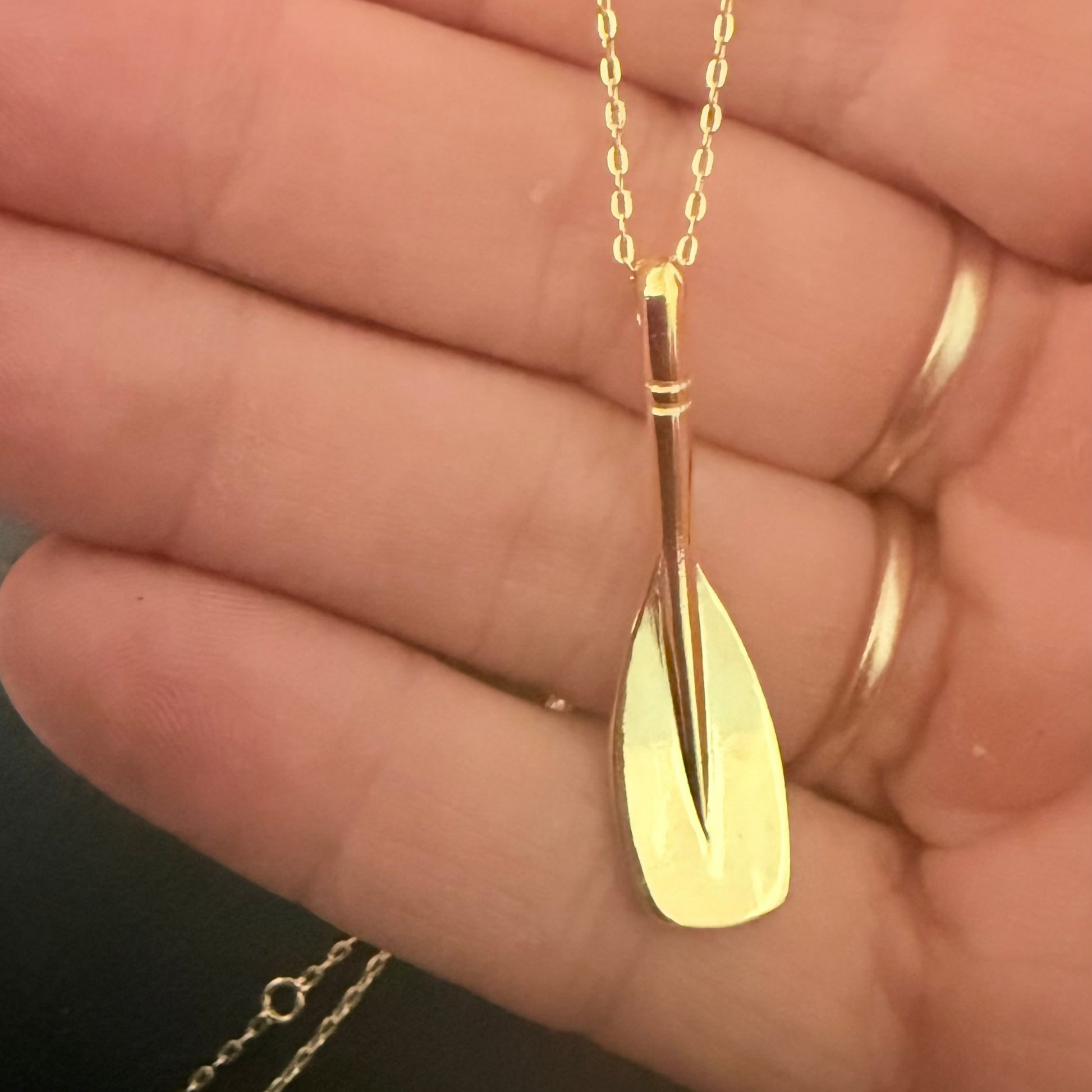 A gold pendant necklace shaped like a stylized oar or rowing blade, hanging from a delicate gold chain and held in a person's hand.