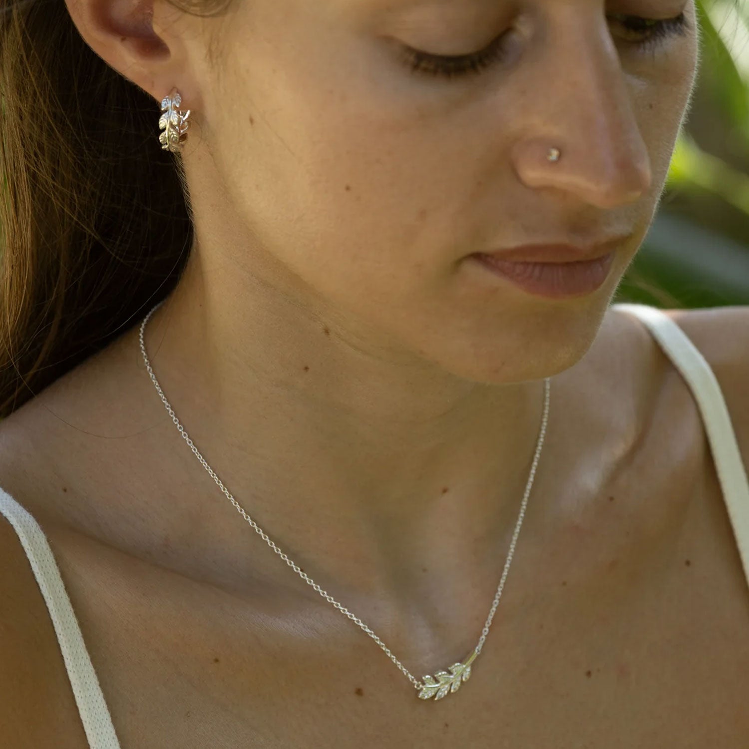 A close-up lifestyle shot of a woman wearing a delicate silver laurel leaf pendant necklace and a matching silver earring.