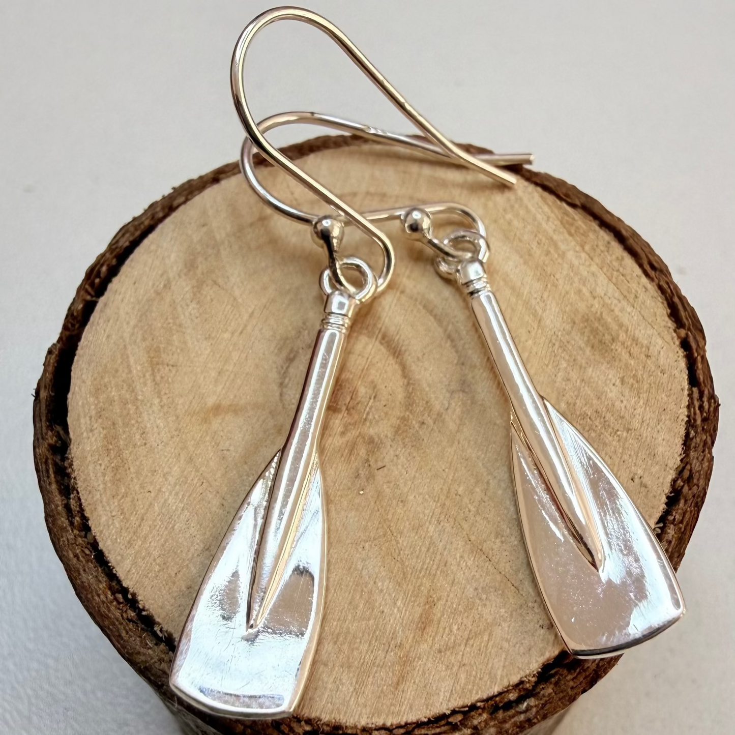 A pair of silver drop earrings shaped like oar blades, displayed on a round wooden slice against a light neutral background.
