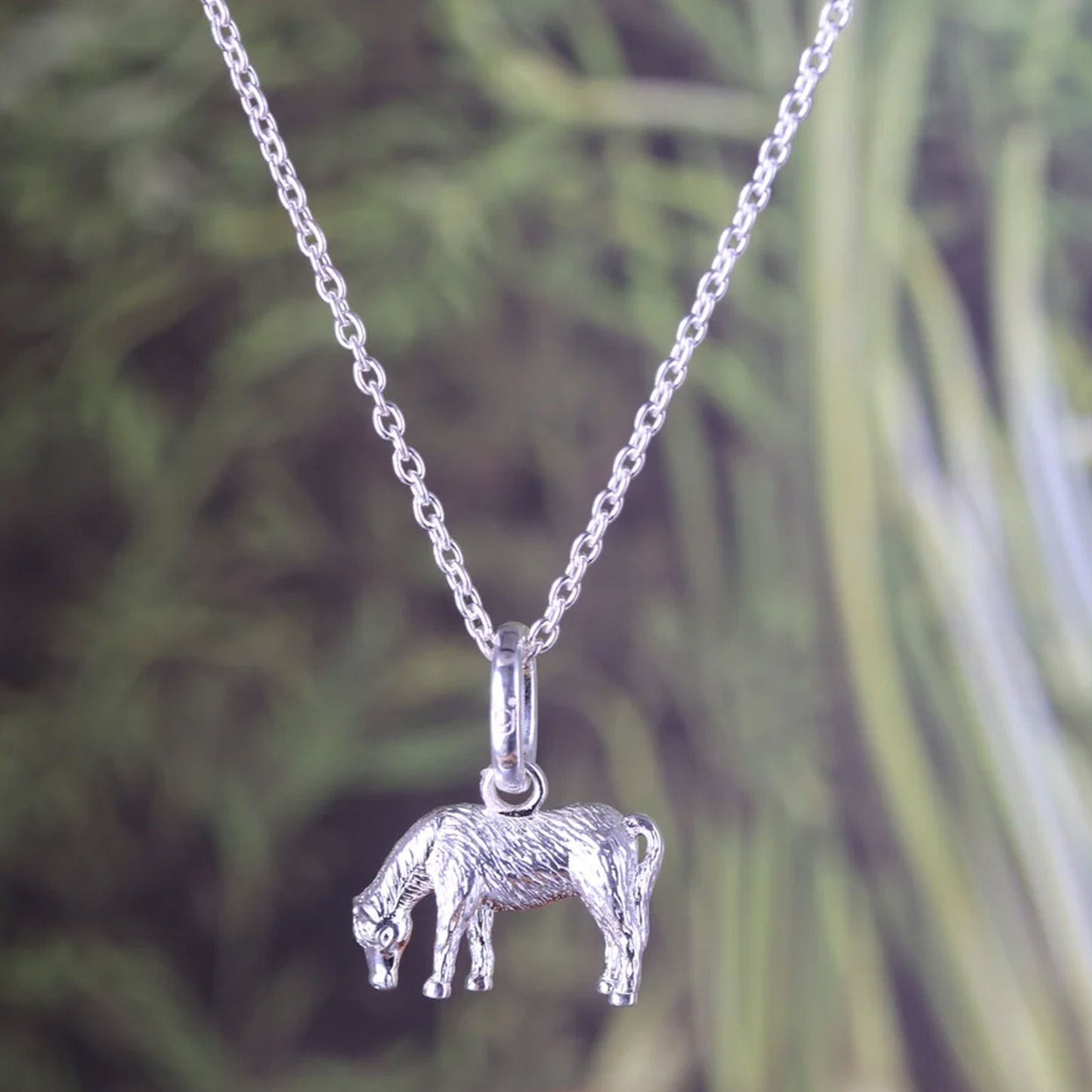 A sterling silver pony pendant necklace on a delicate silver chain, set against a blurred green foliage background.