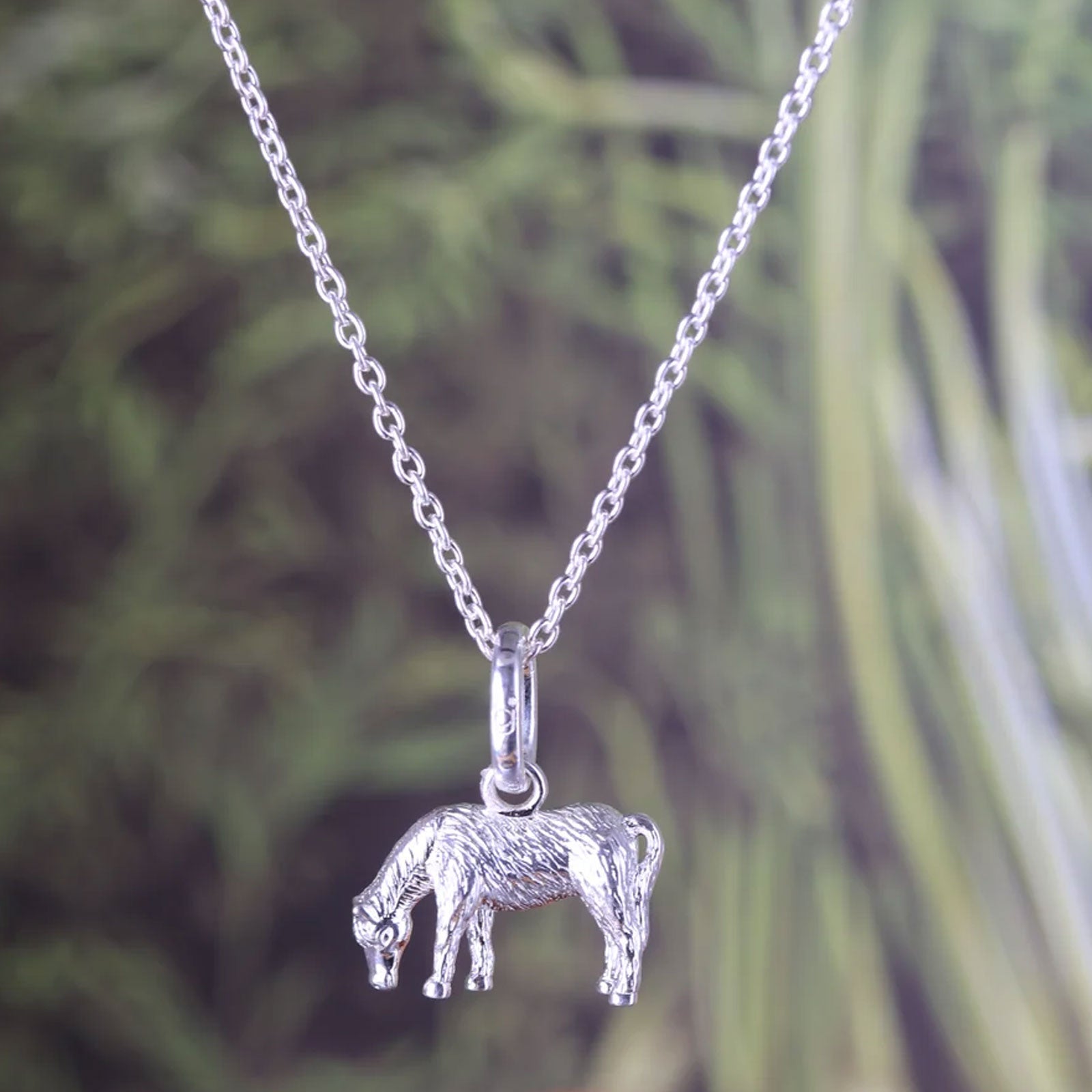 A sterling silver pony pendant necklace on a delicate silver chain, set against a blurred green foliage background.
