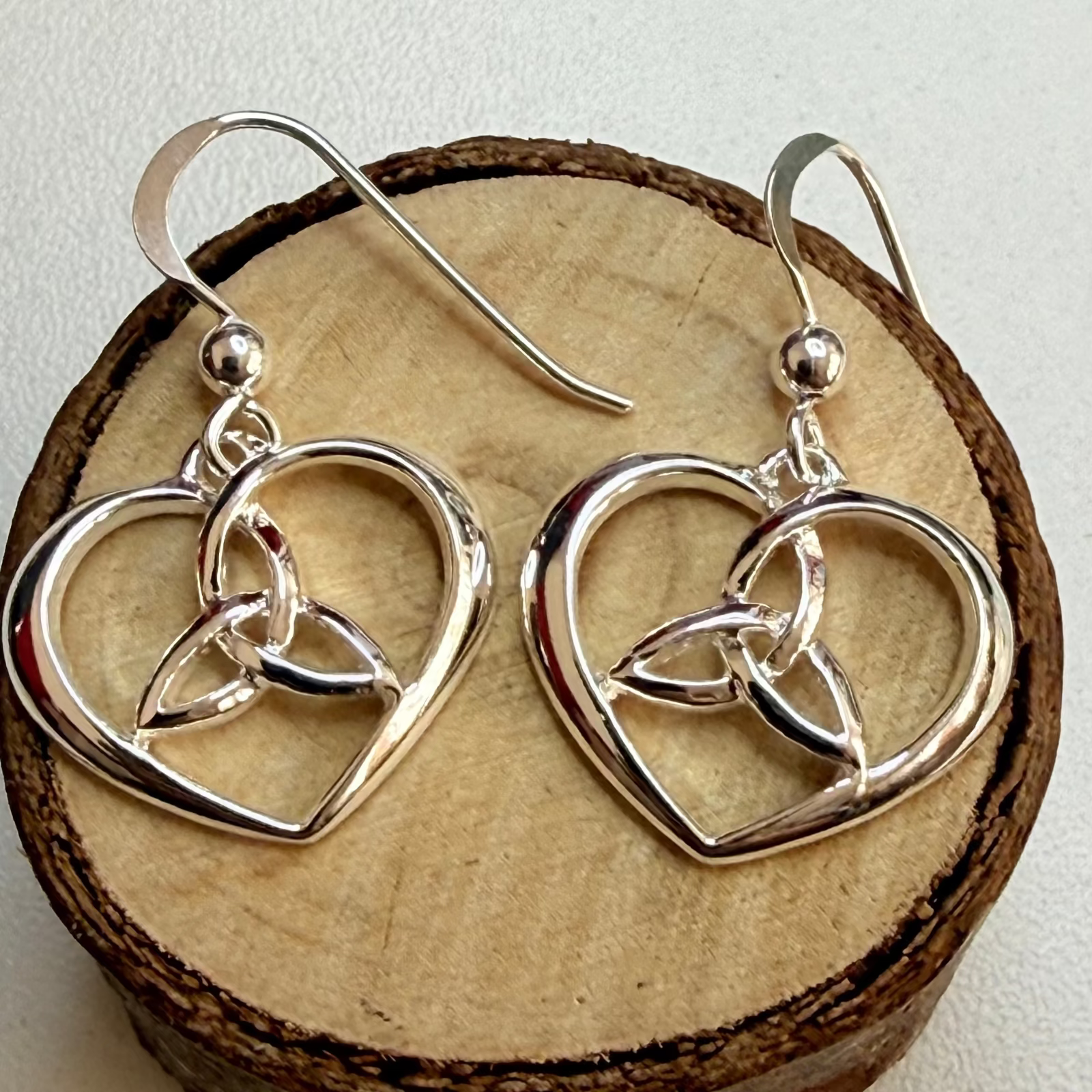 A pair of silver dangle earrings with interlocking heart and Celtic knot designs, displayed on a round wooden slice against a light beige background.