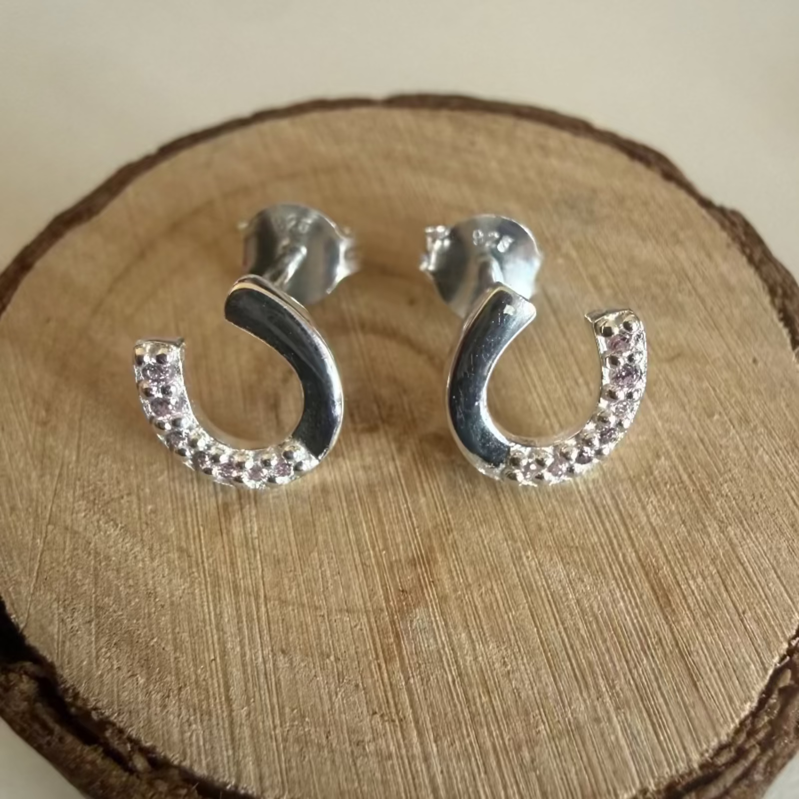 A pair of silver horseshoe earrings with small clear cubic zirconias along one side, displayed on a round wooden slice stand against a light neutral background.