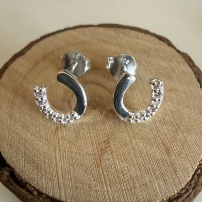A pair of silver horseshoe earrings with small clear cubic zirconias along one side, displayed on a round wooden slice stand against a light neutral background.