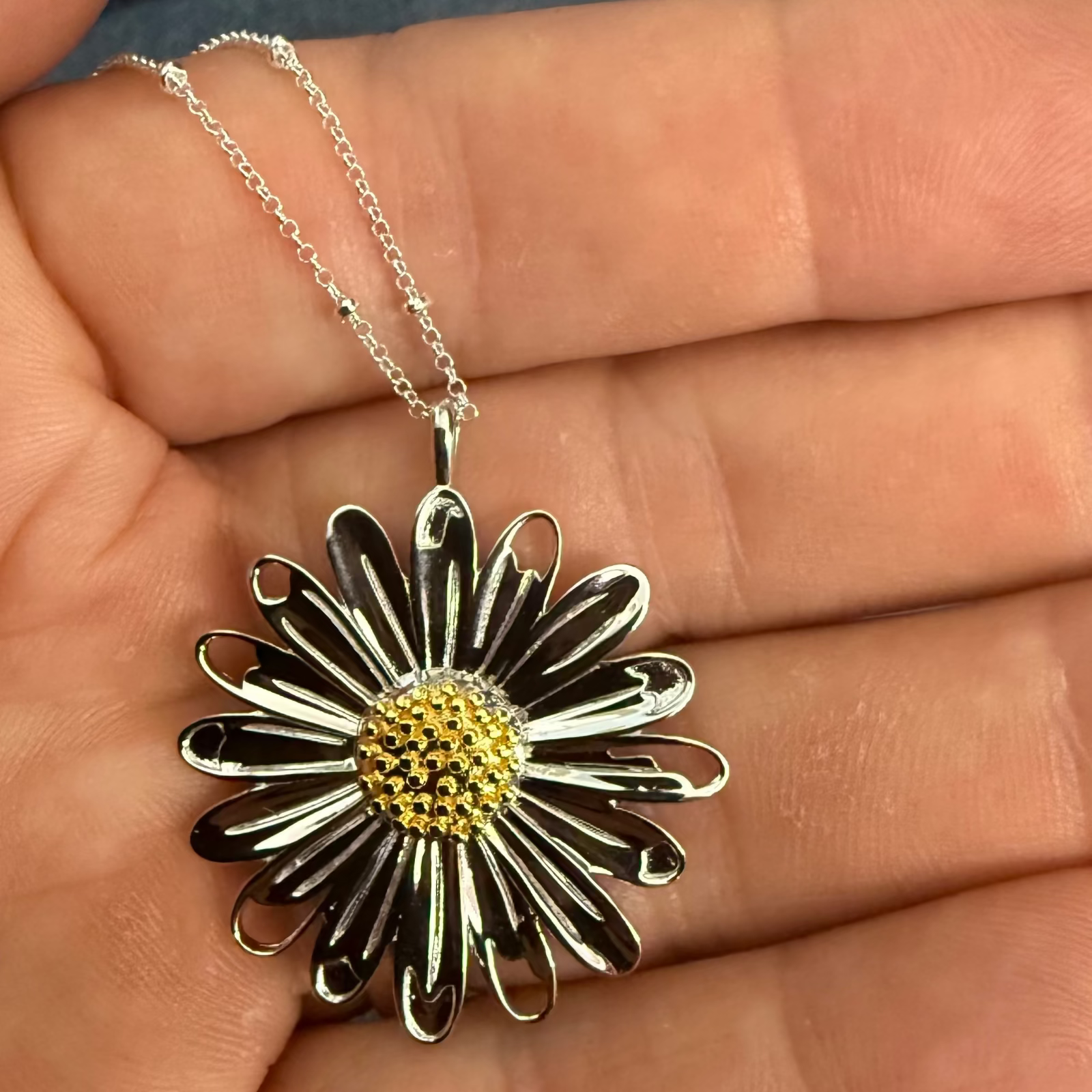 A silver daisy pendant necklace with a textured yellow center, held in a person's hand against a background of blue denim.