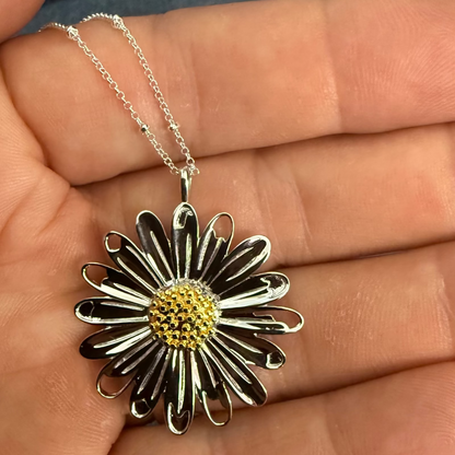 A silver daisy pendant necklace with a textured yellow center, held in a person's hand against a background of blue denim.