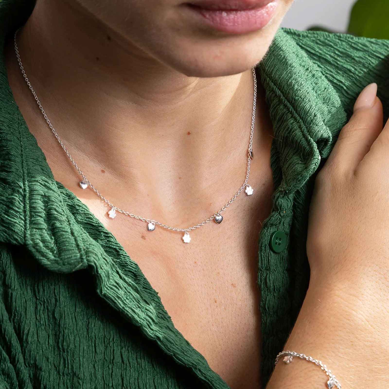 Person wearing a delicate silver necklace with small dog paw and heart charms against a green garment.