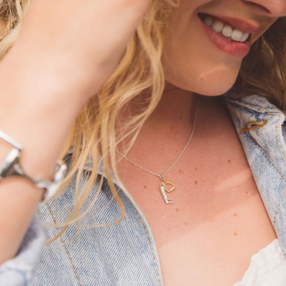 A close-up lifestyle shot of a woman wearing a delicate silver chain necklace with a small gold stirrup and silver boot pendant. She is wearing an open light blue denim shirt, and her blonde hair is visible in the foreground.