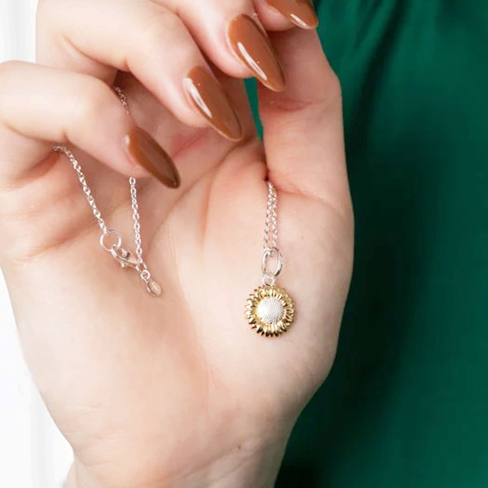 Hand holding a gold and silver necklace with a sunflower pendant against a green background
