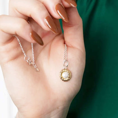 Hand holding a gold and silver necklace with a sunflower pendant against a green background