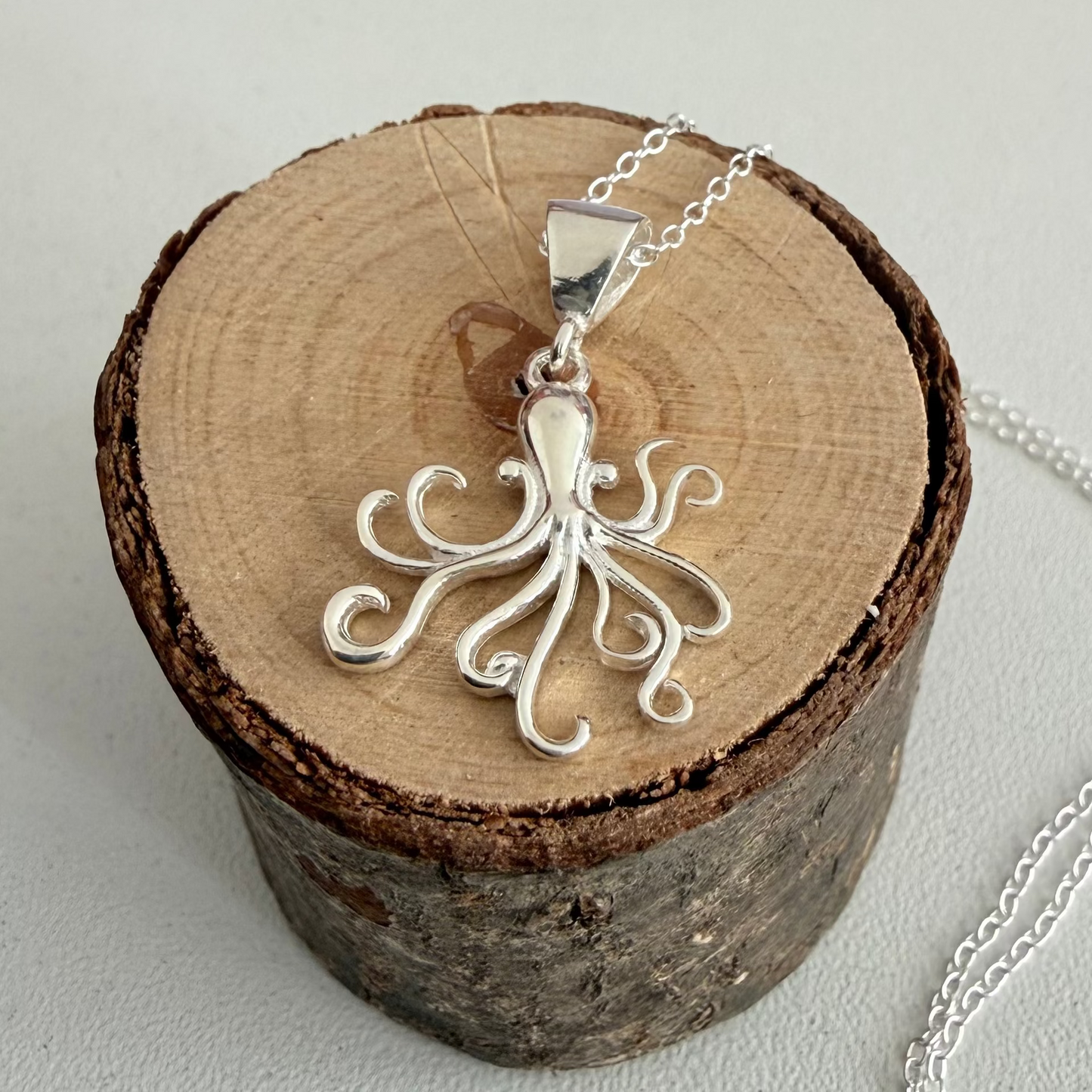 A silver octopus pendant necklace displayed on a round wood slice stand, with part of the silver chain visible on a light gray background.
