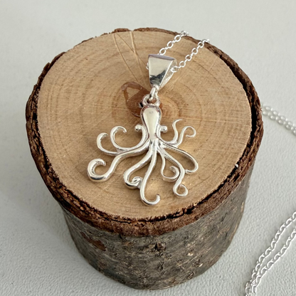 A silver octopus pendant necklace displayed on a round wood slice stand, with part of the silver chain visible on a light gray background.