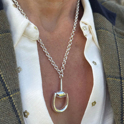 A close-up of a person wearing a silver horse bit pendant necklace over a white collared shirt and a brown plaid blazer.
