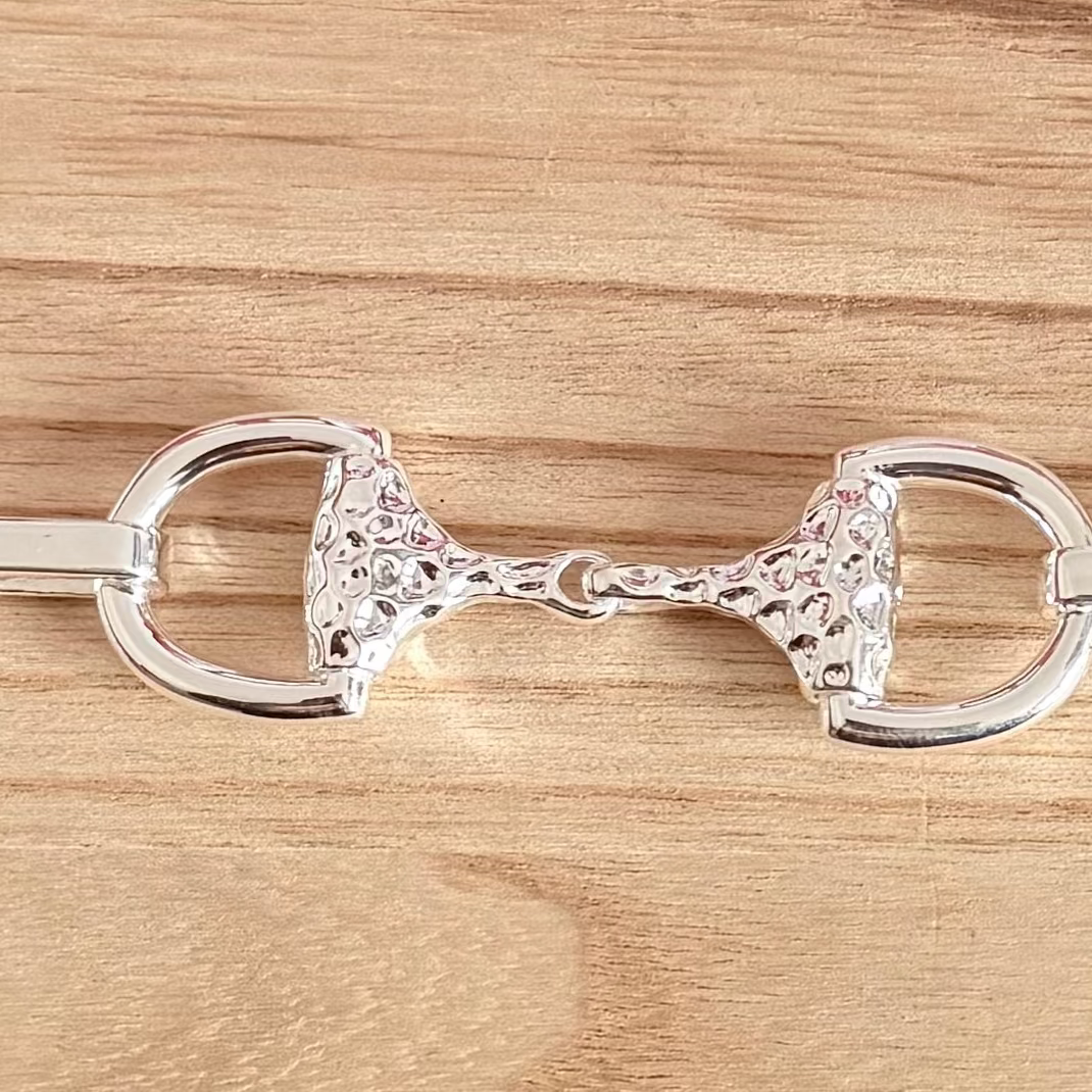 A polished silver horse bit with a textured, hammered pattern on the cheekpieces, displayed horizontally on a light brown wooden surface.