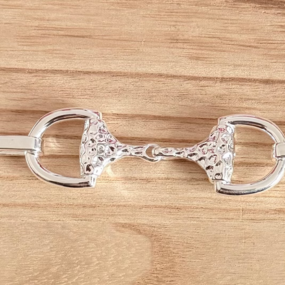A polished silver horse bit with a textured, hammered pattern on the cheekpieces, displayed horizontally on a light brown wooden surface.