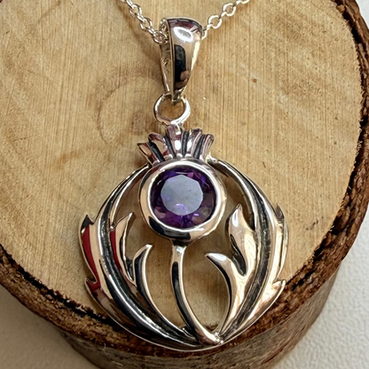 A sterling silver pendant necklace featuring a stylized thistle design with a round purple amethyst gemstone, displayed on a rustic wood slice background.