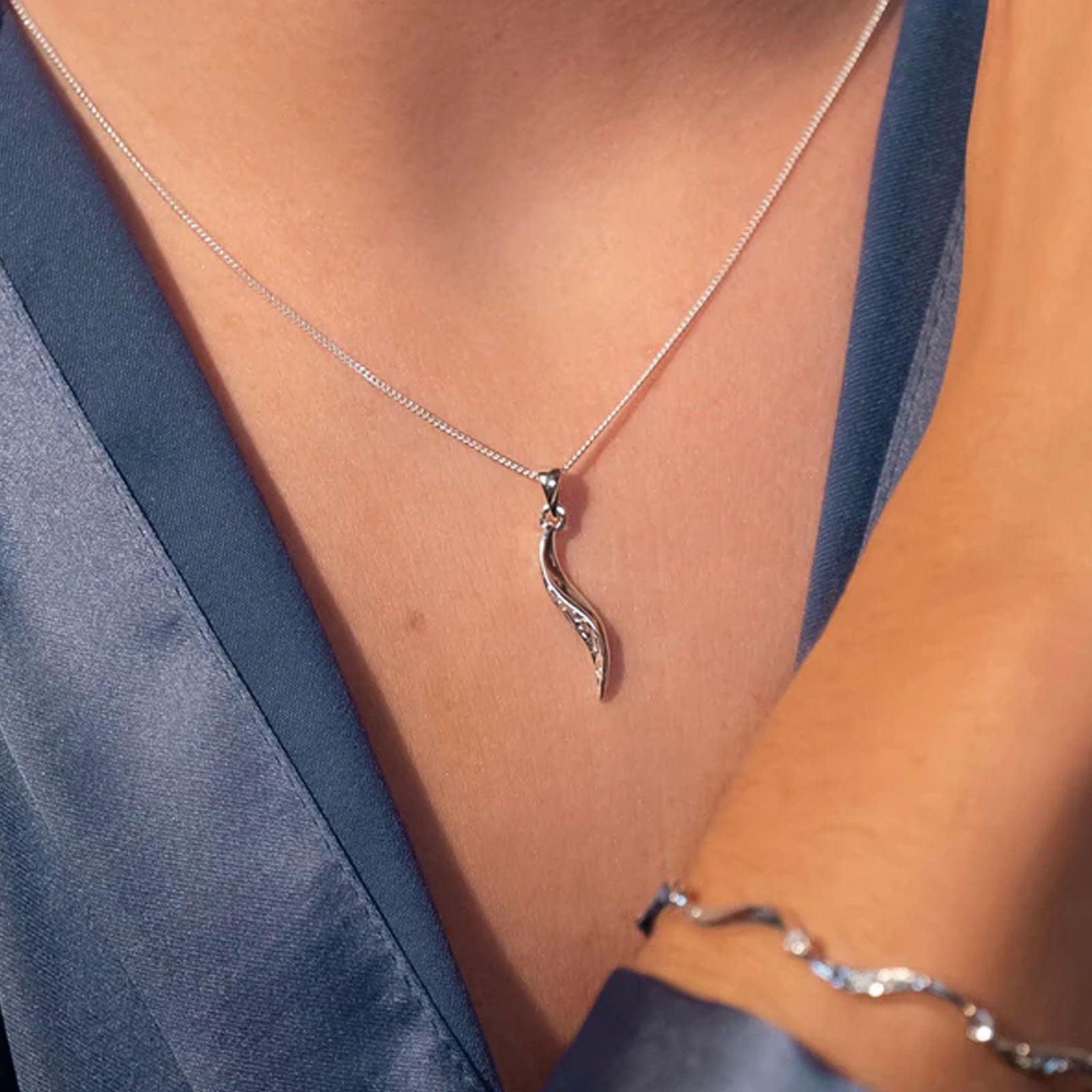 A close-up of a model wearing a silver wavy pendant necklace and a matching silver bracelet, styled with a blue satin robe.