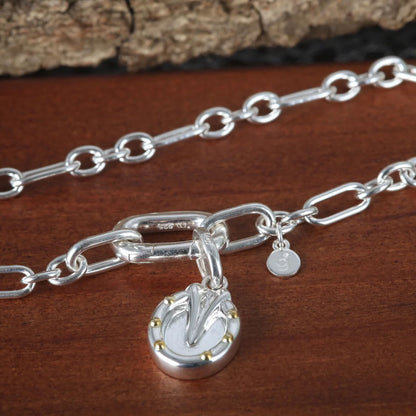 A sterling silver chain necklace with a large oval link clasp, a horseshoe charm engraved with a stylized letter, and a small hanging tag, displayed on a dark wood surface.