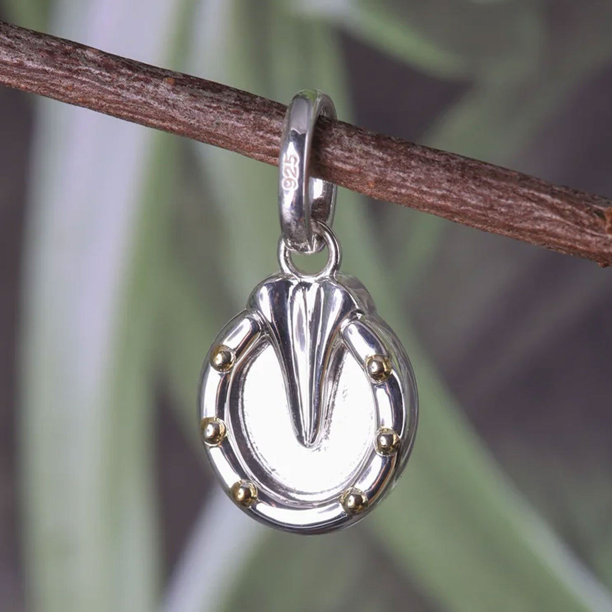 A sterling silver horseshoe charm with gold-colored rivets, hanging from a brown twig against a blurred green and gray background.