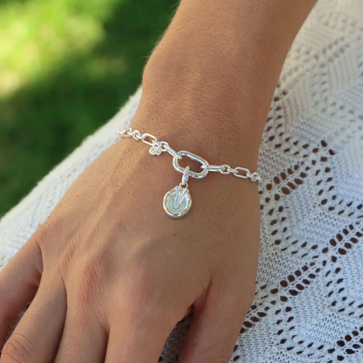 A sterling silver chain bracelet with a well shod horse shoe charm, shown worn on a person's wrist over a white lace top with a blurred green outdoor background.