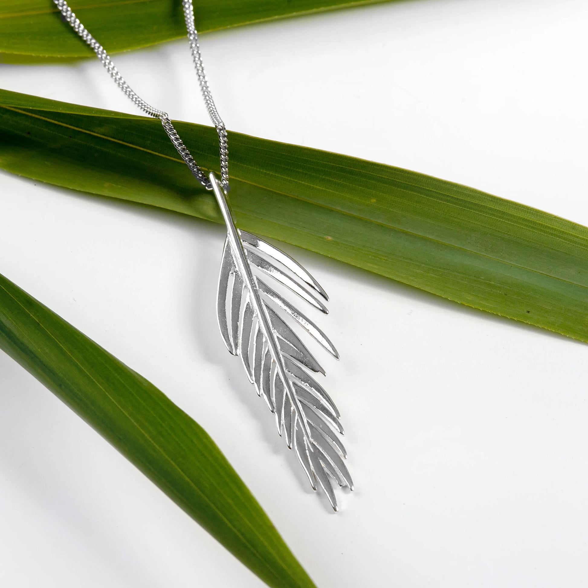 Silver feather-shaped pendant on a chain with green leaves in the background